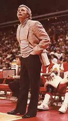 A man with curly gray hair, wearing a tan jacket and shirt with black pants and shoes, standing with his hands in his pockets