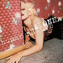 Madonna lying on the ground against a red wall with flowers painted on it. She wears a black dress and her face is half hidden behind the wall.