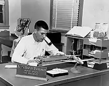 Photograph of a man with a crew cut at work writing, sitting at a desk that has a model fighter plane on it