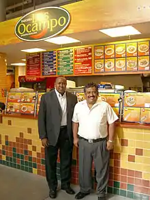 Two men, a taller one on the left in an open suit jacket and the shorter one on the right in a white button-down shirt, smile as they stand in front of an indoor taquería.