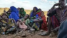 A group of at least 4 children, 4 women and a 1 man sitting on the ground.