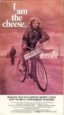 A red-toned scene  shows a teenage boy on a bike, one foot down on the path to keep himself still. His left elbow rests on one handlebar while he grips the other. There is a white package in the basket. He looks thoughtful and possibly confused, his brow lowered. Behind him and on the left side of the cover, a man stands in a dark jacket and pants; his face is not visible. On the right, behind both the man and the boy, is a white farmhouse with a black roof. The sparse trees surrounding it look bald. The cover reads "I am the cheese." in white font. The white square below the red cover lists the credits of the film. Most visible are in black at the top of the white square; it reads: "Robert MacNaughton, Hope Lange, Don Murray and Robert Wagner."