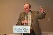 MacPherson, a mature Caucasian man wearing a suit and tie, speaking at a conference. Gesturing with one hand, he stands behind a podium bearing a convention centre logo.