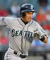 A man in a gray baseball uniform running to the right. The uniform reads "Seattle" across the front, and has a sleeve patch with a teal and silver compass surrounded by "Seattle Mariners". The man is wearing a dark batting helmet.