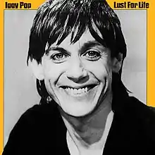 A black-and-white portrait of a middle-aged man with long hair grinning at the camera, with a yellow border. The words "Iggy Pop" appear in the top left corner; the words "Lust for Life" appear in the top right.