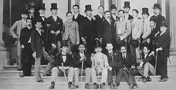 Photo of a large group of men on steps. Some are seated, and others are standing; several are wearing top hats.