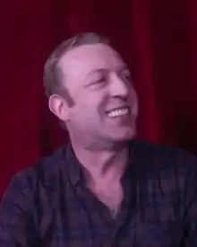 Neidell smiling against a red curtain, facing right