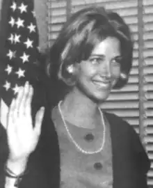 A smiling young blond woman, wearing a dark jacket over a sheath-style dress, stands in front of a window and an American flag, with one hand raised