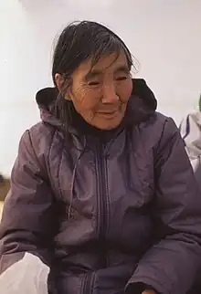 An older woman sits outdoors. She has a medium skin tone and long black-and-grey hair, and is wearing a purple parka.