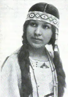 A young woman with tan skin and dark hair in two long braids, wearing a beaded headband and a leather tunic with beading around the neckline