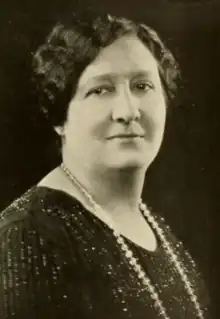 A white woman with dark hair, wearing a dark dress and a strand of beads or pearls