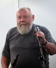 Ivo Pešák wearing a grey t-shirt with breast pocket, holding onto a microphone in a stand with one hand, with a clarinet in the other, looking directly at camera