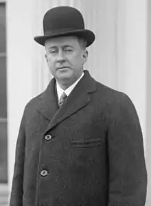 Cleanshaven man with drooping eyelids, aged about 40. He is wearing a black bowler hat, white shirt, tie and dark overcoat.