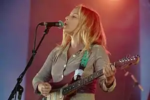 Upper body shot of a 27-year-old woman. She is singing into a microphone with her head slightly tilted up and is seen in mostly left profile. She is also playing an electric guitar. The artist is wearing a champagne coloured top, which is sparkling. Her fair-to-blonde hair is over her shoulders. The background has alternate panels of purple-blue and plum sections.