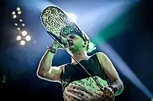 White male standing onstage, holding microphone to his mouth, wearing tank top and unusual hat with brim