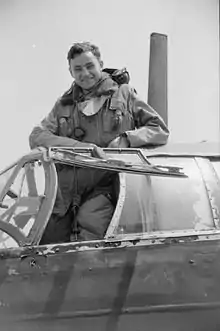 A black and white photograph of a man in a flying suit standing in the cockpit of an aircraft