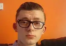 Photographic portrait of a white teenage boy with slicked-back brown hair. He is wearing a chequered white shirt and black, thick-framed glasses. He is sitting on a black leather settee in front of an orange wall.