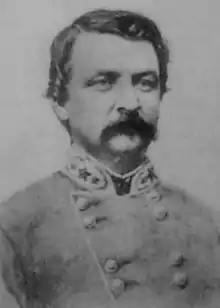 Black and white photo shows a man with a moustache wearing a gray military uniform with two rows of buttons. The insignia on the collar indicates a Confederate general officer.