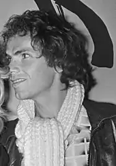 A black-and-white photo of a young Dutch man with lengthy dark-colored hair. He is wearing a faux leather jacket, a pattern shirt, and a scarf. He smiles as he looks to the left of the camera.