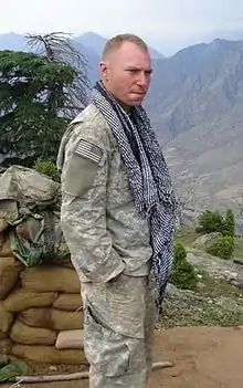 A man in his camoflouge military uniform wearing a scarf around his neck. He is looking at the camera and has his hands in his pockets. Several mountains are visible in the background.