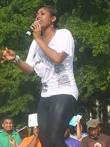 A female with long, straight, black hair poses, wearing a button-up ensemble outfit and donning gold earrings.