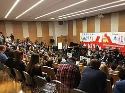 People in a lecture hall observe a group of musicians with instruments as a teacher speaks into a microphone