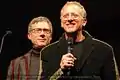 Rob Epstein holding a microphone and smiling, while Jeffrey Friedman is seen standing behind him. Both wearing glasses and facing to the front