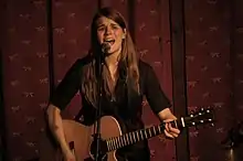 Woman playing acoustic guitar and singing into a microphone in front of a red-patterned background.