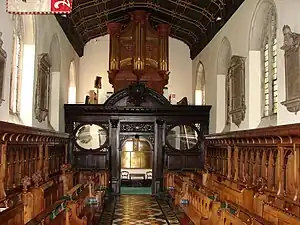 An organ with gilded pipes on top of a decorated wooden screen, with two rows of wooden benches on either side of an aisle