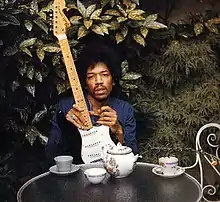 A color image of a man sitting at a table in the outdoors holding a black guitar.