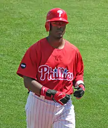 A dark-skinned man in a red baseball jersey, white baseball pants with red pinstripes, and a red batting helmet walks on a baseball field while taking off his black and red batting gloves.