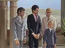 Three men and a boy stand in a palatial setting. A desert landscape is visible from a balcony in the background. One man on the far left is grey-haired and wears a grey suit and tie, the man to the right of him dark-haired and in deep navy blue. Both men are orientated in the direction of the (blond-haired) boy, who is also formally attired in grey. The third man, also blond but wearing a cream-shaded suit, stands directly behind him.