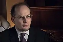 A man with a short beard and glasses, wearing a suit.