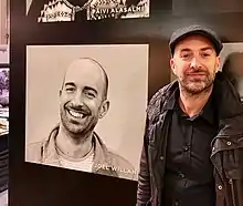 Color photograph of author Joel Willans standing next to a portrait of himself at a book fair booth