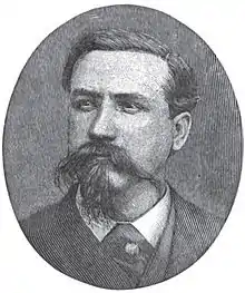 A man with dark hair and a dark, wiry mustache and goatee wearing a white shirt and black jacket