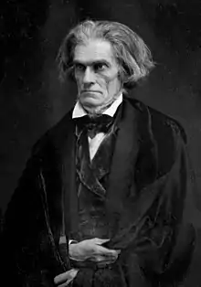 Age 67, long gray hair, austere look, frowning, holding black cloak closed with both hands.