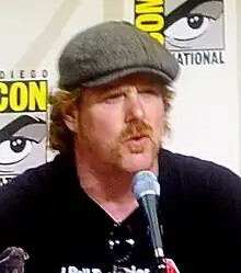 A bearded man wearing a black shirt and a gray beret sits in front a microphone. Sunglasses are han