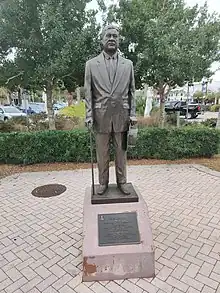 Statue of elderly man in suit and tie, holding a brimmed hat and a walking stick..