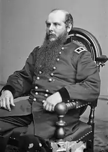 Black and white photo of a seated man in a dark military uniform with the shoulder tabs of a major general (two stars). He is balding and has a curly beard that reaches halfway to his waist.