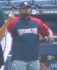 A bearded, dark-skinned man in a red-and-navy blue jacket and gray baseball pants throwing a baseball with his right hand