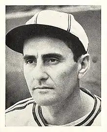 A man in a white baseball jersey and cap with dark stripes looks–the left.