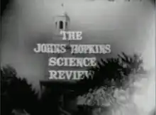 Television screenshot looking up at the bell tower of a building through tall trees. The  words "The Johns Hopkins Science Review" are superposed on the image.