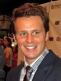 Young Caucasian man with short, blonde hair smiling and looking away from the camera. He is wearing a suit, collared shirt and tie.