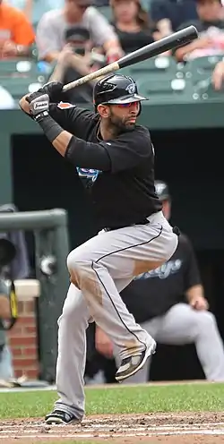 A man in a black batting helmet, black baseball jersey, and grey pants holds a baseball bat midway into right-handed swing with his left leg up in the air.