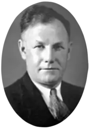 A head and shoulders, black and white photographic portrait of a white man with receding dark hair