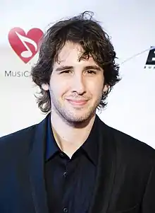 A young man with medium-length wavy hair, smiling slightly and dressed in black business attired.