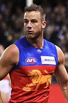 A man in a maroon, blue and gold football jersey and maroon shorts jogs on grass