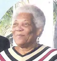 Image of an older woman with a grey afro, she is wearing a v necked striped dress or shirt