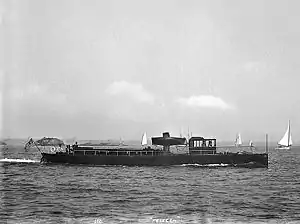 An image showing a ship at sea in calm conditions. The ship is short and long with a singular flag being flown at the rear. Four white sailing ships are shown behind and to the right of the ship in the far background.