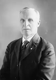 Black and white photo. Man with full white mustache and short hair wearing dark unbuttoned jacket with dark dotted necktie